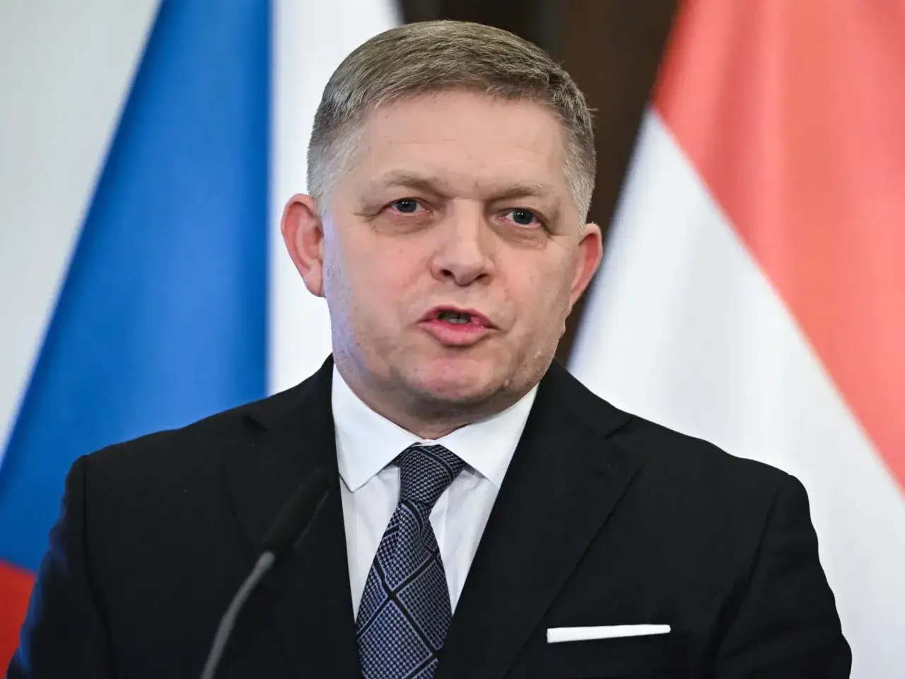 Premier Słowacji w garniturze i krawacie, przemawia na tle flag.