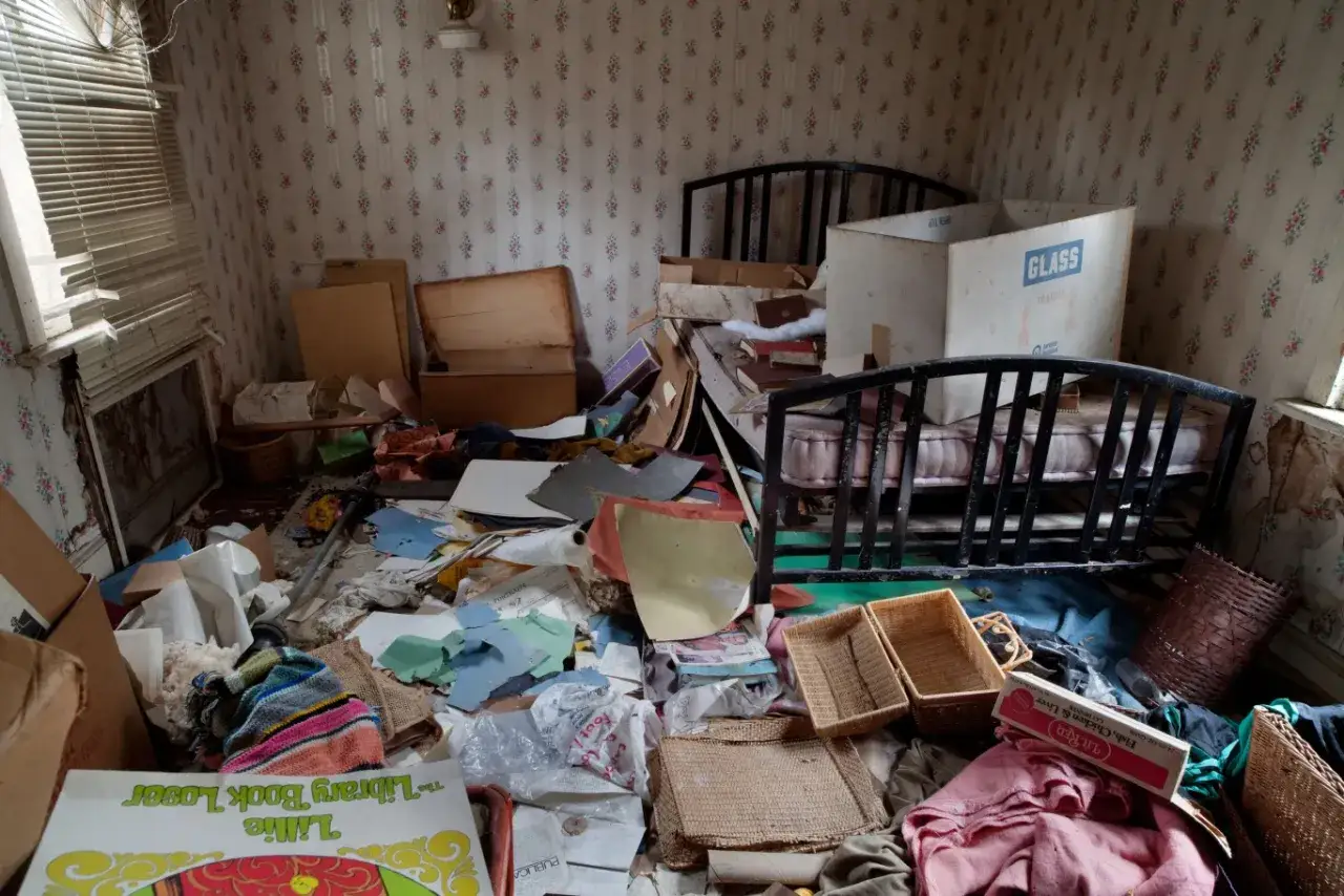 Pokój w stylu Diogenesa, pełen śmieci, starych mebli i papierów. Widać łóżko, skrzynie i kartony.