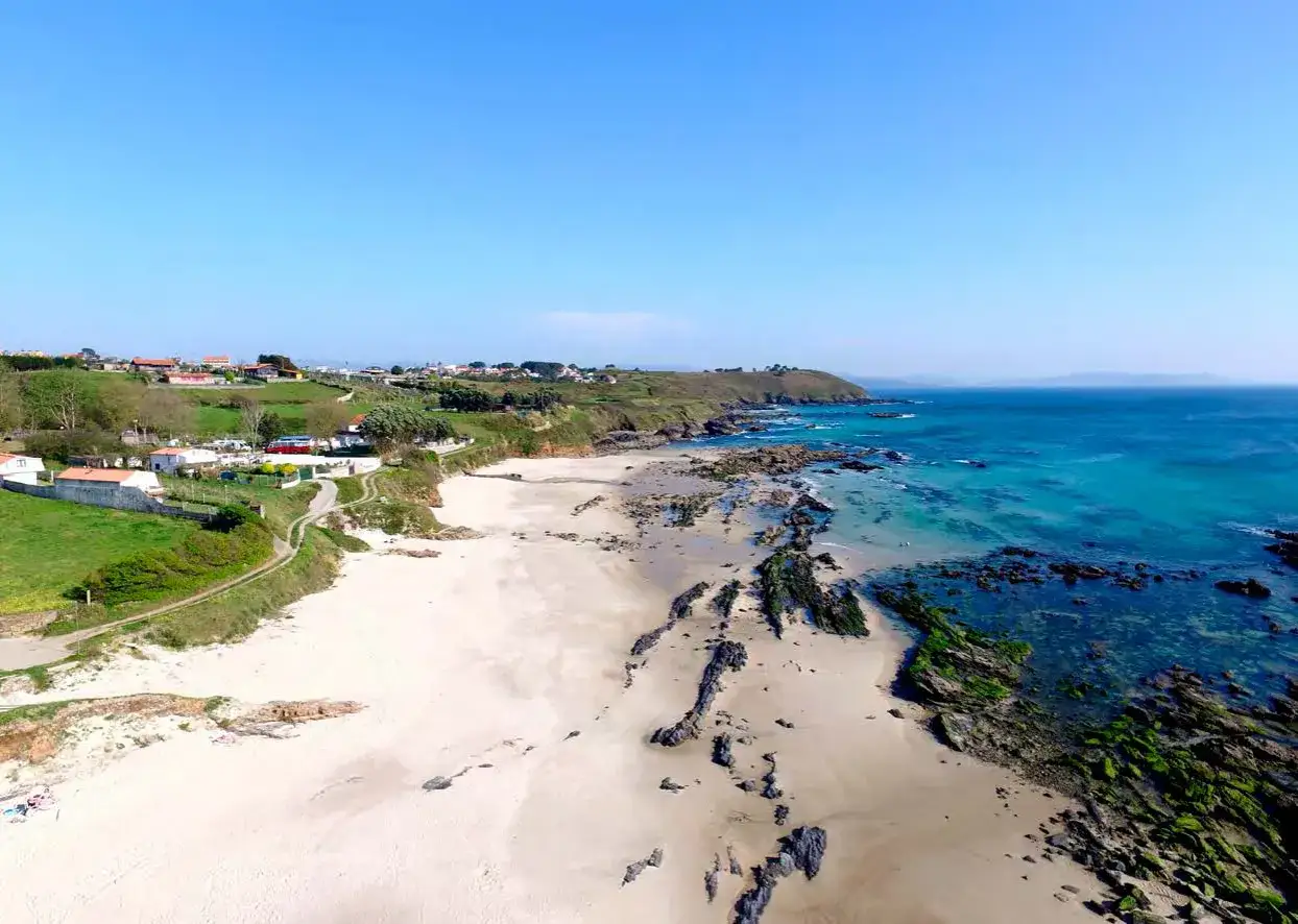 Vista aérea de la playa pragueira, con arena clara, rocas cubiertas de algas y el mar azul turquesa. Al fondo, un pueblo costero y colinas verdes.