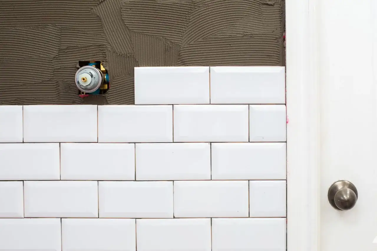 White subway tile installation in progress, with exposed plumbing fixture and a door frame visible.
