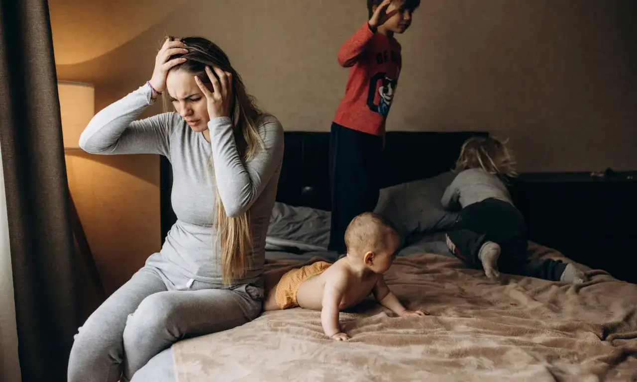Een moeder met handen op haar hoofd, overweldigd door de chaos van haar kinderen. De dynamiek van adhd en autisme in een gezin.