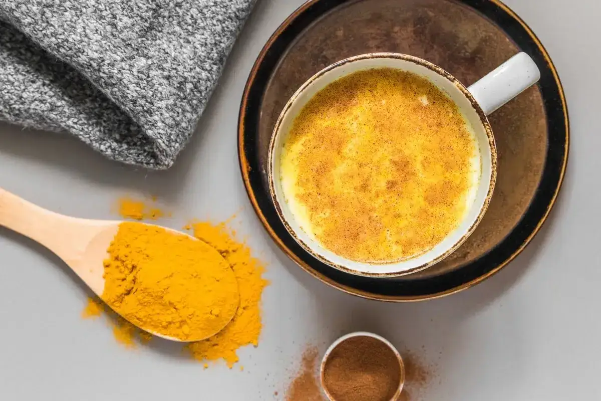 Une tasse de lait au curcuma, avec une cuillère de poudre de curcuma et de cannelle à côté.