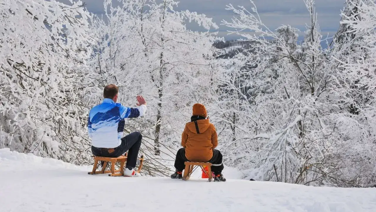 Vater und Kind genießen das Rodeln in Bayern. Verschneite Bäume bilden eine winterliche Kulisse.