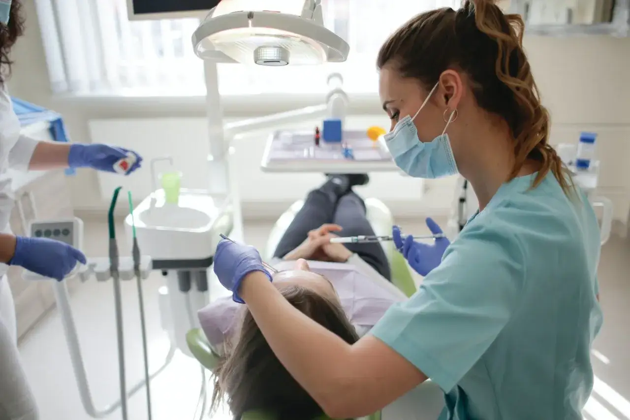 Dentista con mascherina esegue un trattamento. La paziente è sdraiata, con le mani incrociate. Nessuno pensa a fumare dopo pulizia denti.