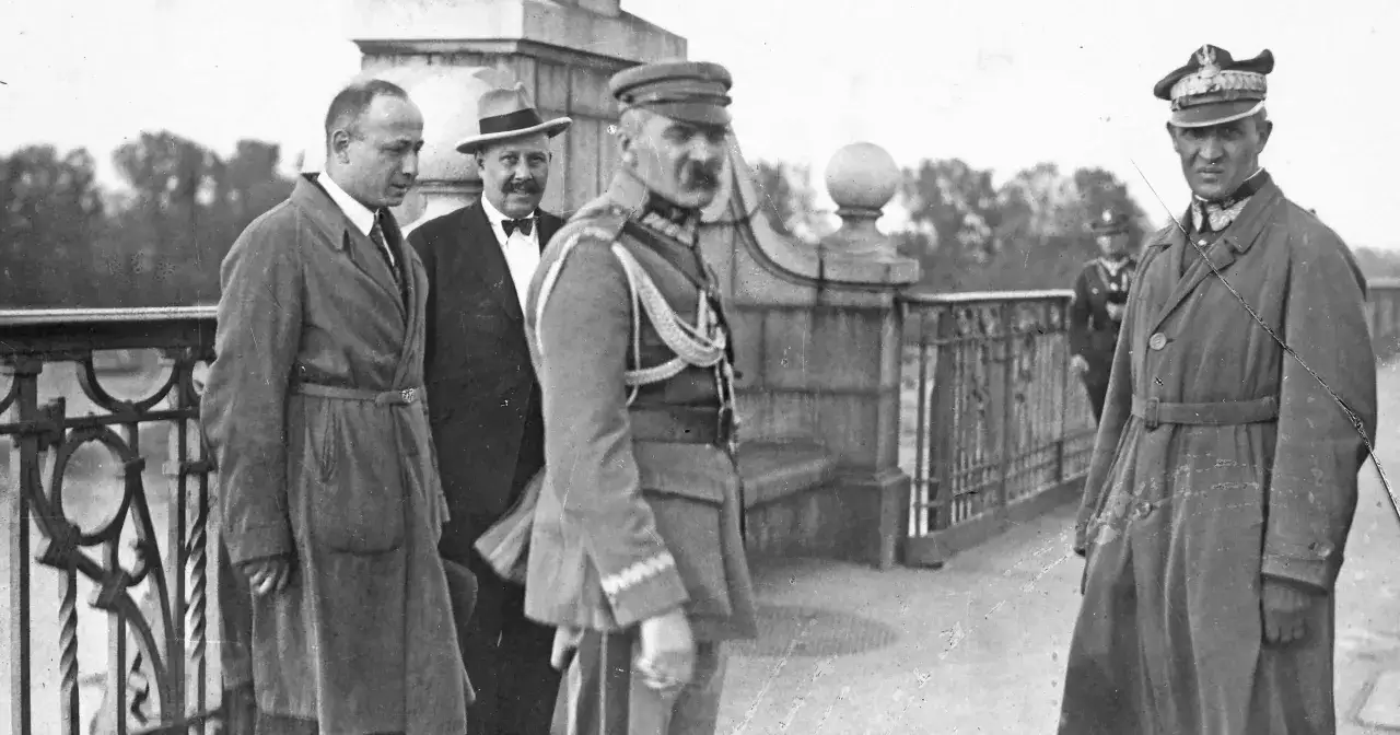 Na moście spotkanie zwolenników Piłsudskiego. W centrum oficer w mundurze, po bokach panowie w płaszczach i kapeluszu.