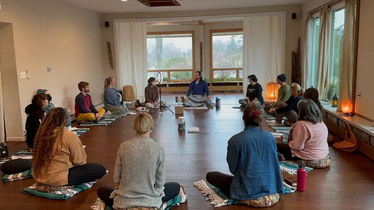 Grupo practicando ahimsa yoga en círculo, meditando en un estudio luminoso con grandes ventanales.