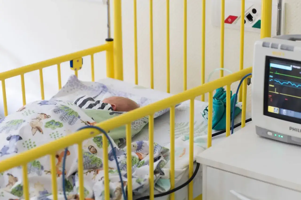 Noworodek śpi w żółtym łóżeczku szpitalnym, otoczony kolorową pościelą. Obok stoi monitor funkcji życiowych, czuwający nad jego stanem.