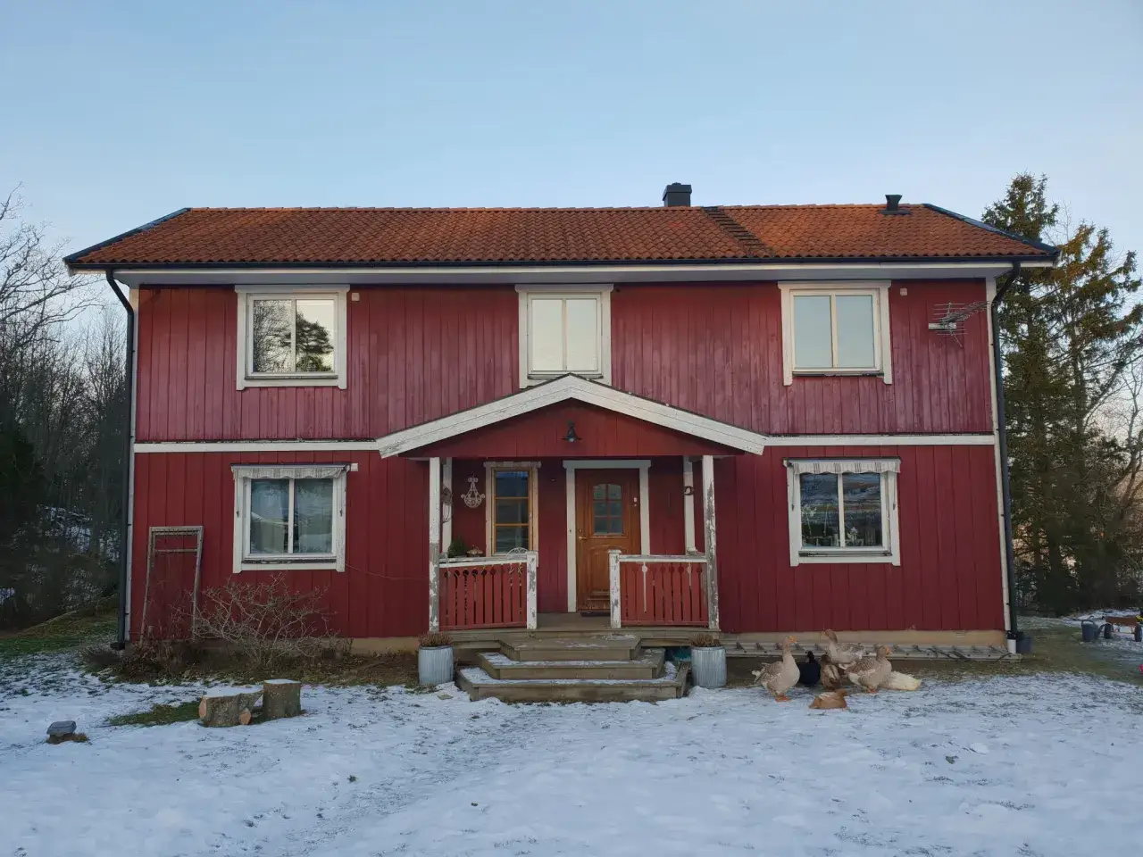 R&ouml;tt hus med tegeltak och en veranda. Fasaden har en fin detalj d&auml;r man lyckats kombinera liggande och st&aring;ende panel. G&auml;ss str&ouml;var i sn&ouml;n.