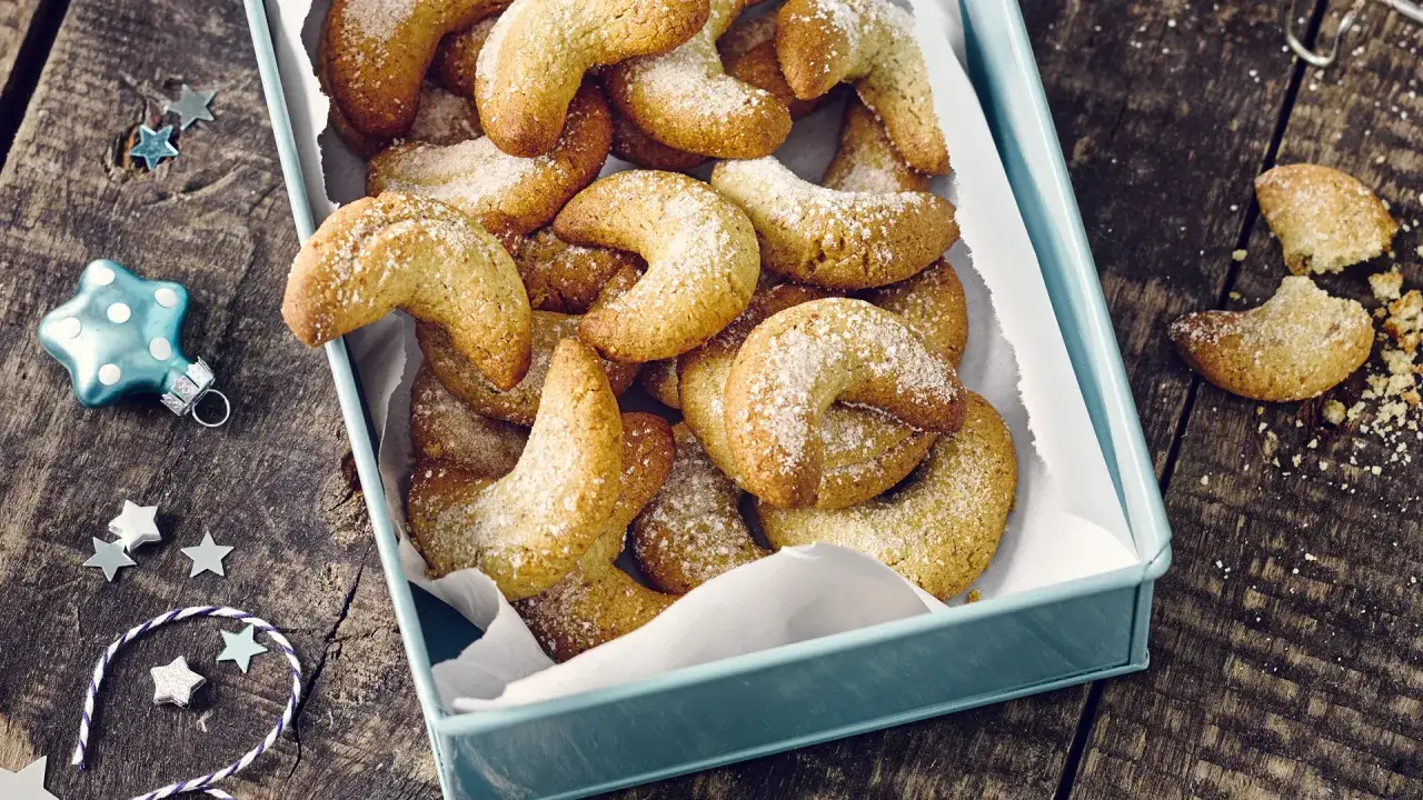 Ein Blech voller goldbrauner Vanillekipferl, bestäubt mit Puderzucker, liegt auf einem rustikalen Holztisch. Weihnachtliche Deko wie Sterne und eine blaue Christbaumkugel schmücken die Szene.