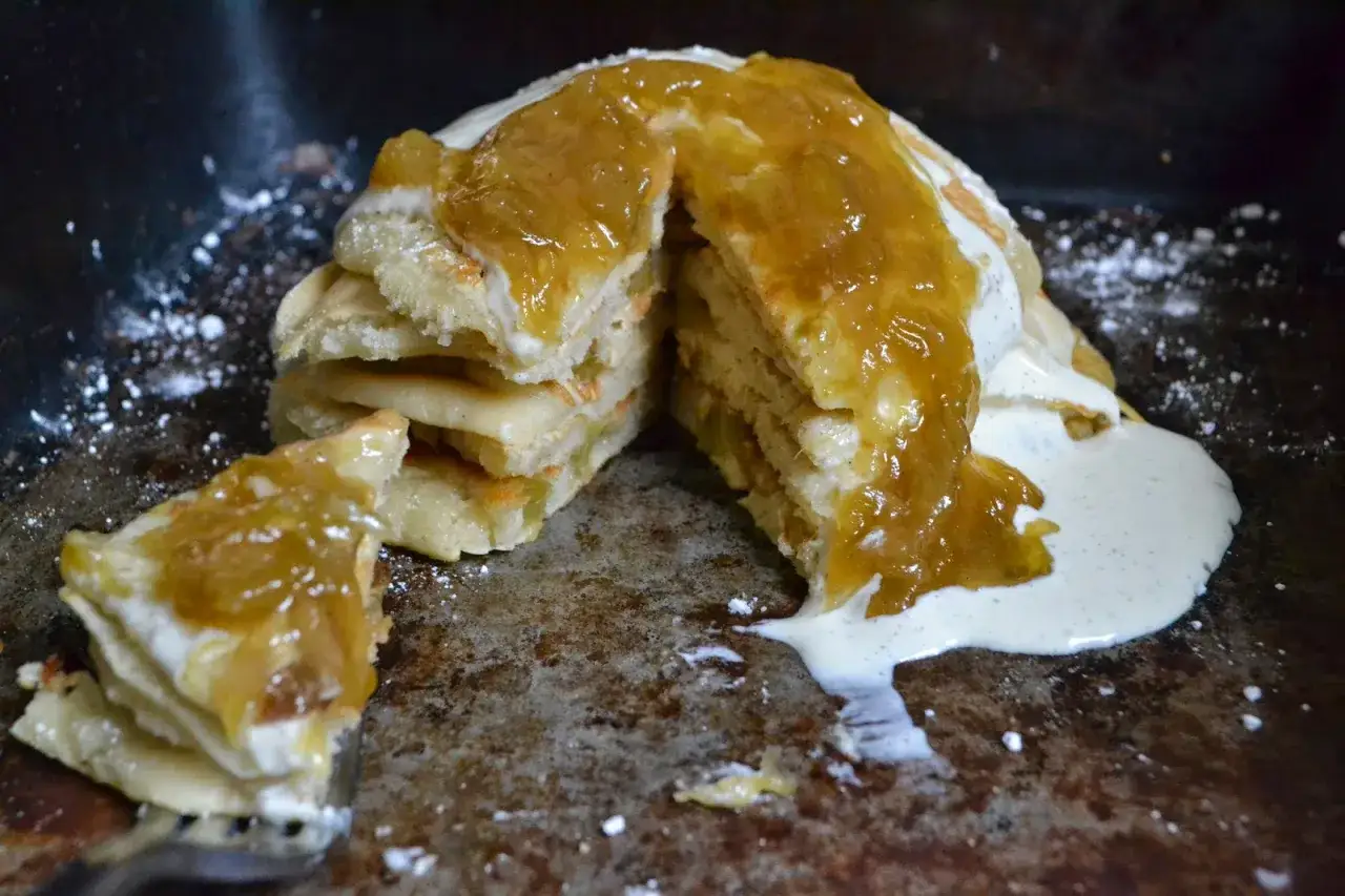 Gestapelte Rhabarber-Pfannkuchen mit cremiger So&szlig;e und Rhabarberkompott, best&auml;ubt mit Puderzucker.