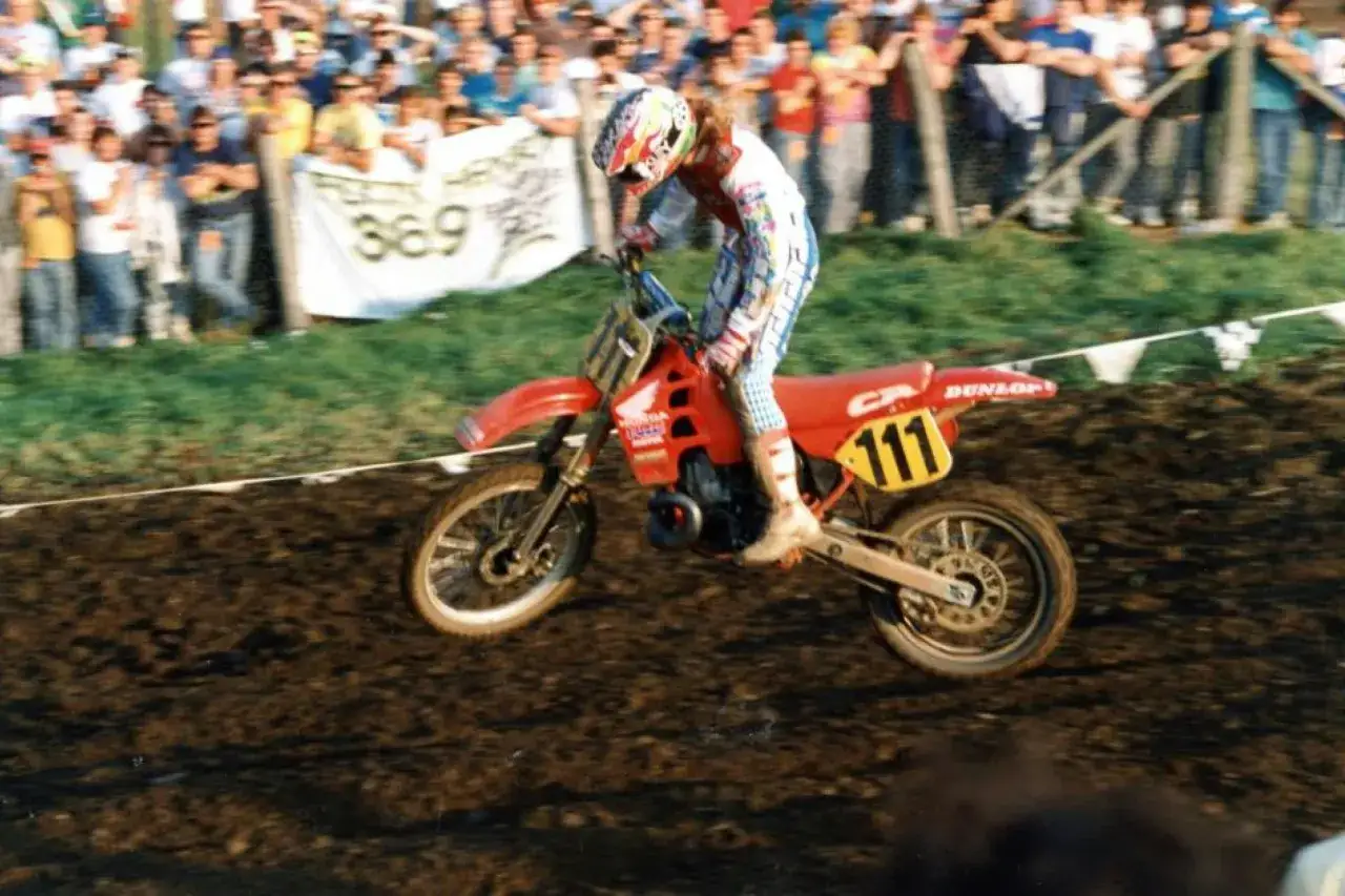 Jean Michel Bayle, con su icónico casco, compite en una carrera de motocross, levantando barro con su moto Honda número 111.