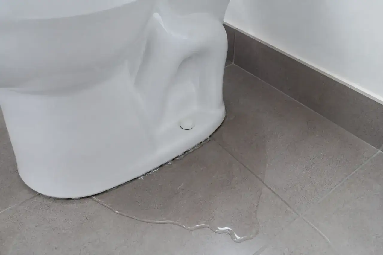 Widok z bliska na białą ceramikę, gdzie sedes przecieka przy podłodze. Woda rozlewa się na szare płytki.