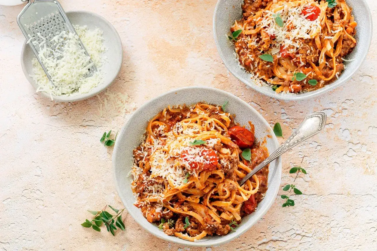 Dwa talerze z makaronem tagliatelle w sosie bolognese, posypane parmezanem. Obok miseczka z tartym serem i tarka. Idealny spaghetti bolognese przepis oryginalny.