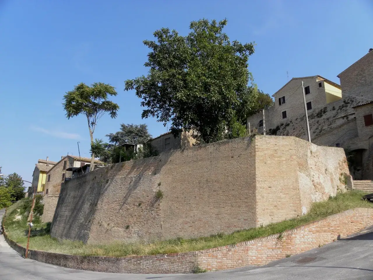 Mura antiche e case in mattoni a Castelleone di Suasa, con alberi verdi e cielo azzurro.
