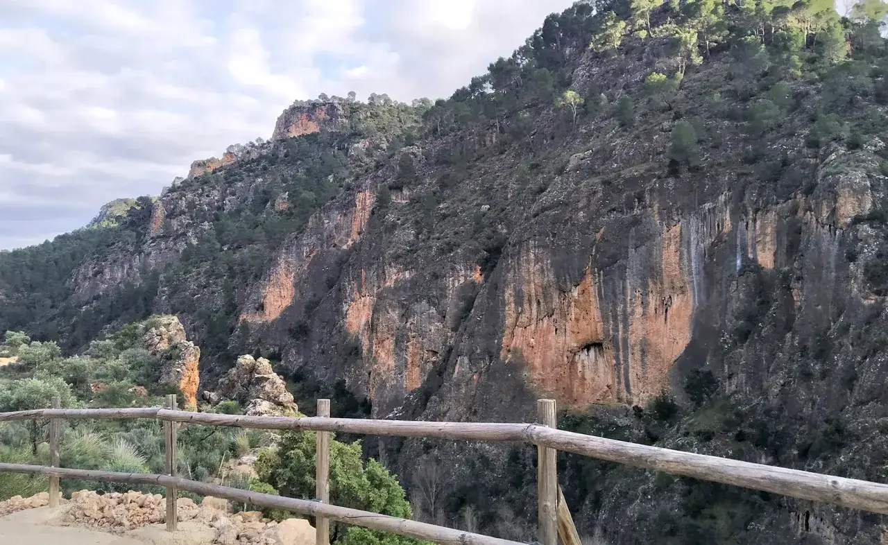 Impresionante pared rocosa con tonos ocres y grises, cubierta de vegetaci&oacute;n. Un mirador del infierno con barandilla de madera ofrece vistas espectaculares.