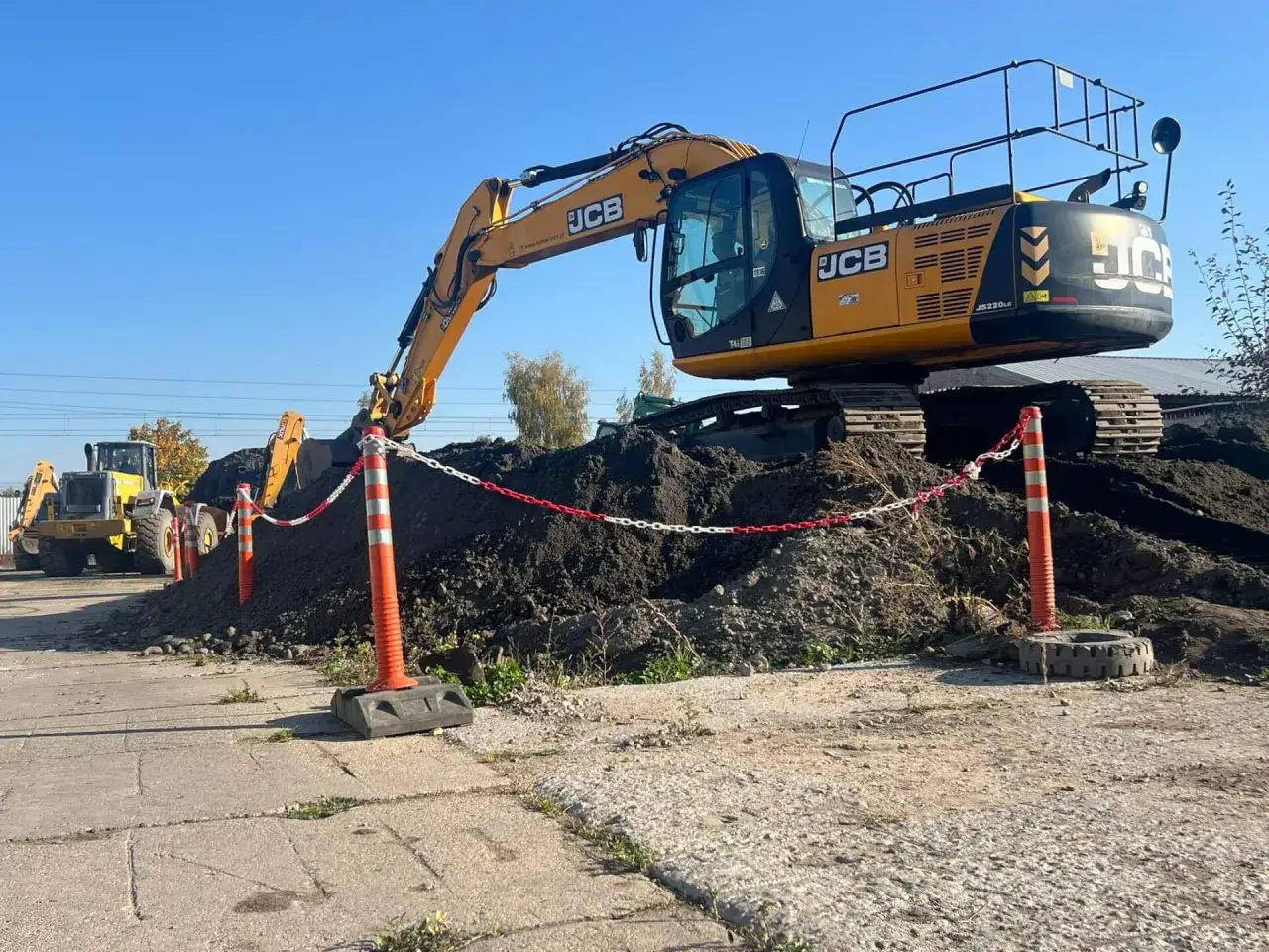 Żółta koparka JCB na placu budowy. Zastanawiasz się, ile kosztuje kurs na koparkę? Maszyna pracuje przy dużych hałdach ziemi.