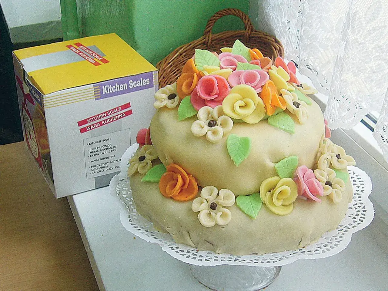 Dwupiętrowy tort z marcepanu ozdobiony kolorowymi kwiatami i listkami. Obok stoi pudełko z wagą kuchenną, która może pomóc w przygotowaniu idealnego marcepanu.