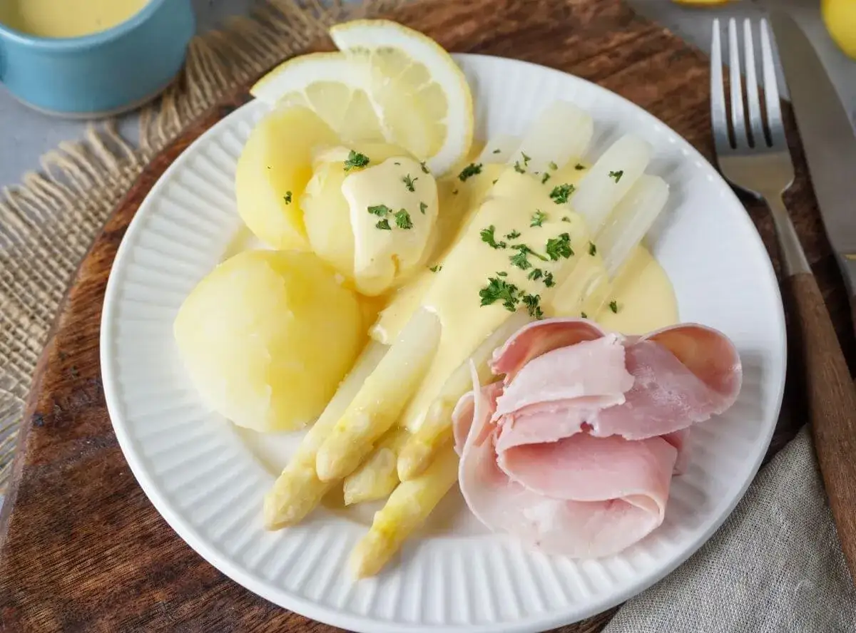 Spargel in Schinken eingerollt, mit Kartoffeln, Sauce Hollandaise und Zitrone auf einem Teller angerichtet.