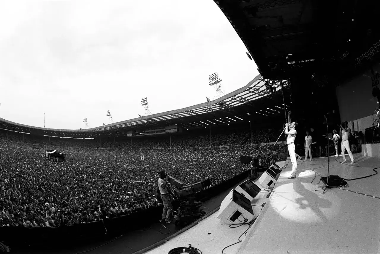 Freddie Mercury śpiewa na scenie podczas Live Aid, tłum wypełnia stadion.