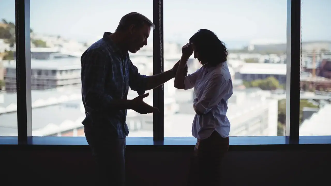 Silueta de pareja discutiendo frente a una ventana. Él le habla con gestos, ella se lleva las manos a la cabeza. Una escena sobre cómo actuar cuando te hacen daño.
