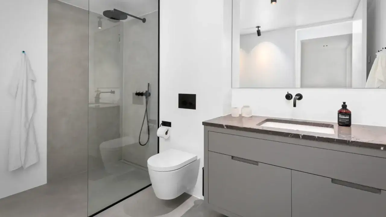 Baño moderno con ducha de microcemento, inodoro suspendido y lavabo con mueble gris. Toalla blanca colgada.