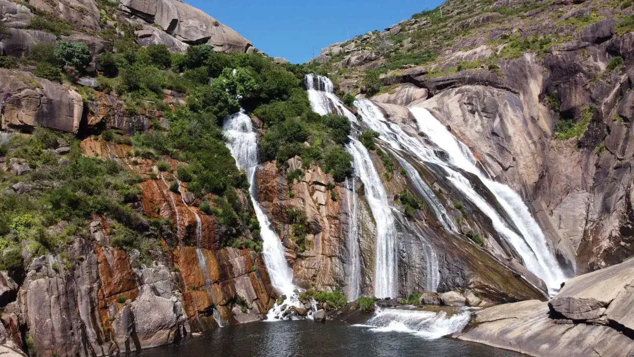 Impresionante cascada fervenza cayendo por rocas rojizas y verdes, creando un espectáculo natural bajo un cielo azul.