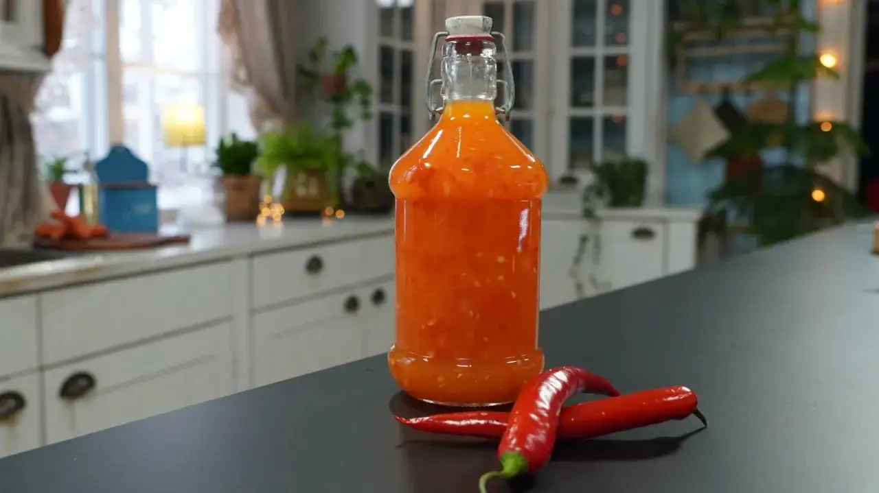 En flaska med hemgjord sweet chili sås står på en mörk bänk, med två röda chilifrukter framför.