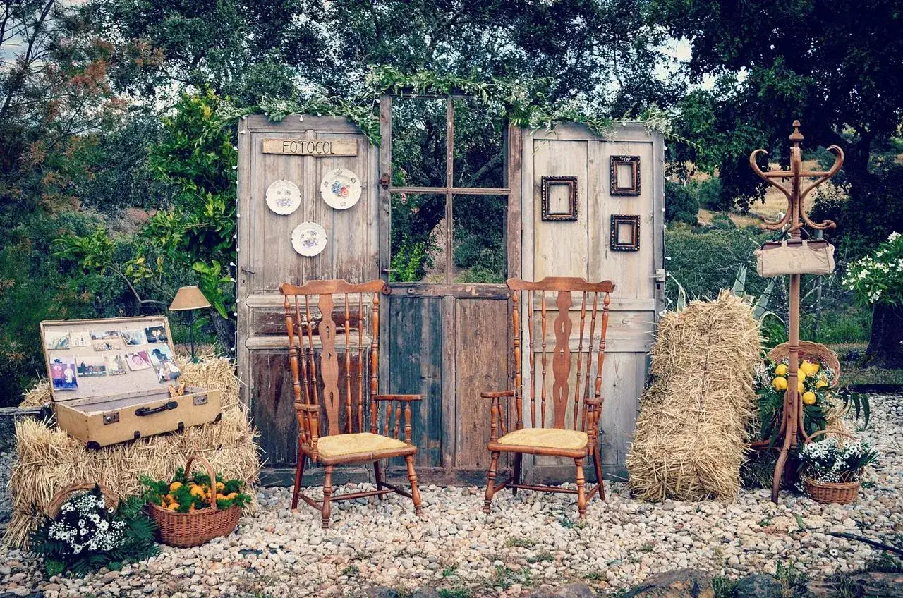 Un photocall bodas de oro casero con puertas r&uacute;sticas, sillas de madera, balas de heno y un ba&uacute;l lleno de recuerdos.