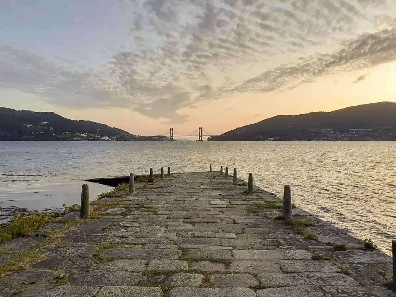 Muelle de piedra en la isla de San Sim&oacute;n al atardecer, con un puente al fondo y monta&ntilde;as cubiertas de vegetaci&oacute;n.
