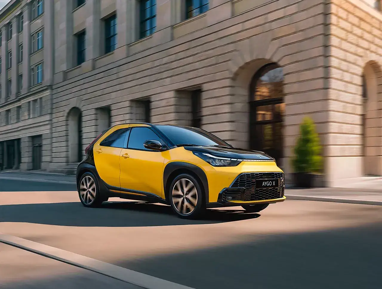 Żółty Aygo X sunie ulicą, mijając kamienną fasadę. Idealny do miejskiej zielonej strefy.