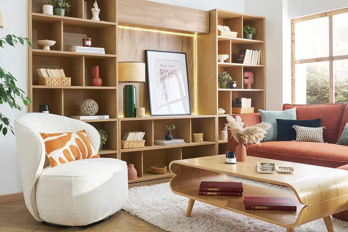 Am&eacute;nagement d'une biblioth&egrave;que dans un salon : &eacute;tag&egrave;res en bois remplies de livres, vases, et objets d&eacute;co, avec un fauteuil blanc et un canap&eacute; orange.