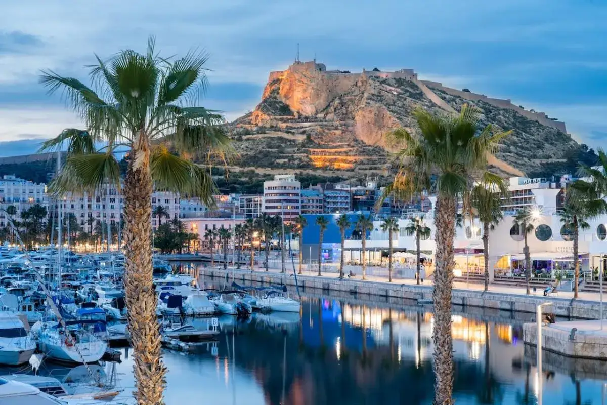 Palme e barche nel porto di Alicante, con il castello illuminato sulla collina al tramonto.