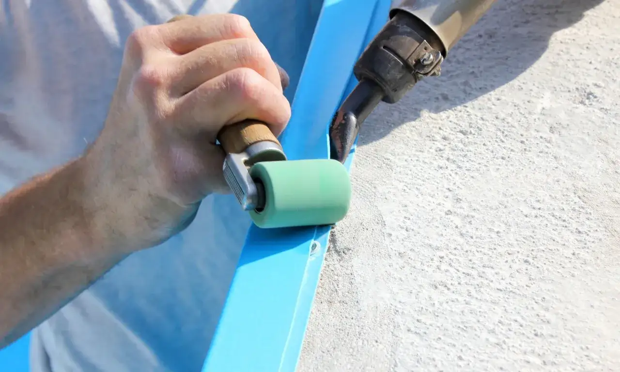 A hand uses a roller to smooth a bright blue material, possibly starboard, as a heat gun melts it to a textured surface.