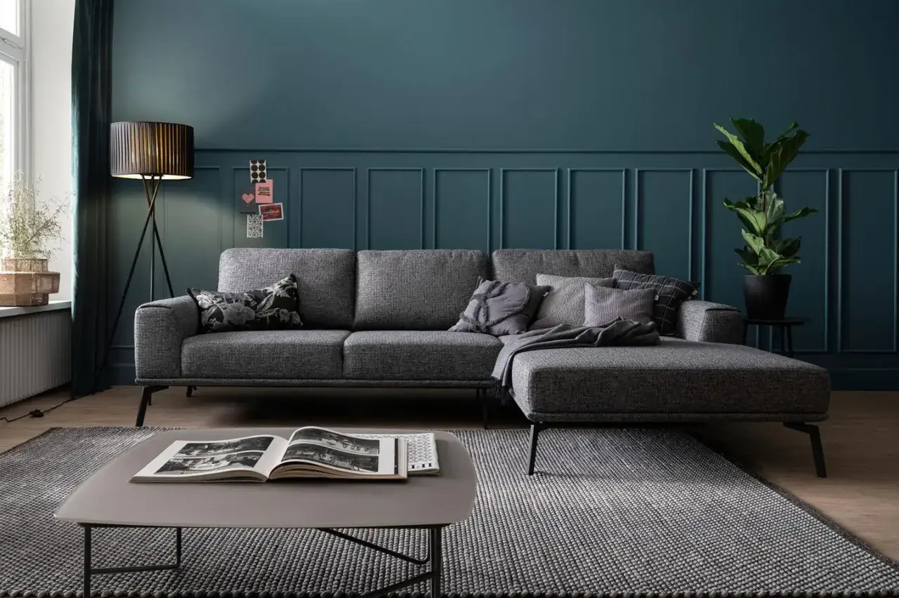 Ein moderner Wohnbereich mit einem grauen Ecksofa, das auf einem gemusterten Teppich steht. Kissen und eine Decke sorgen für Gemütlichkeit.