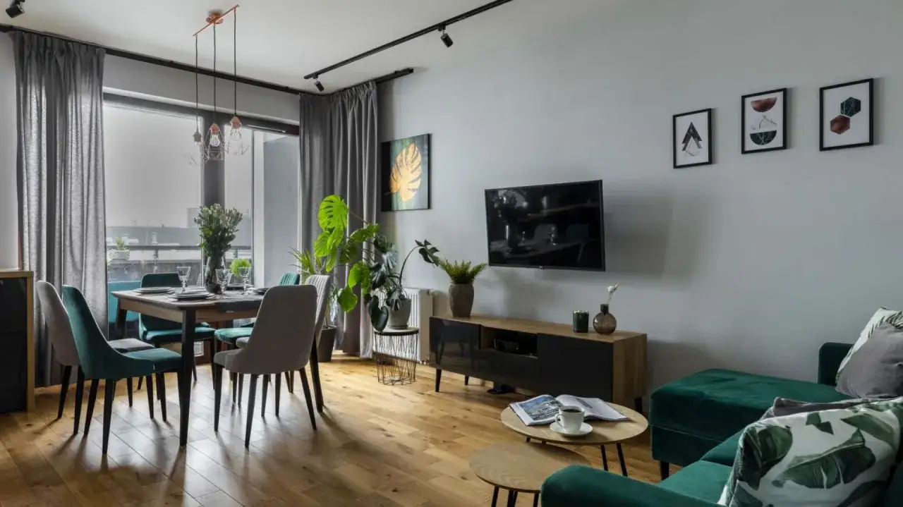 Nowoczesny szary salon z jadalnią. Wygodne, welurowe fotele i kanapa w głębokiej zieleni tworzą przytulną atmosferę. Drewniana podłoga i subtelne dekoracje dopełniają całość.