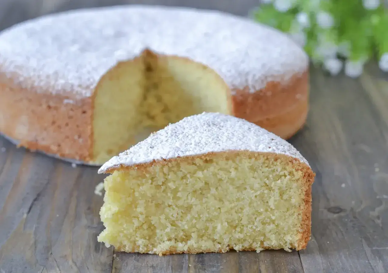 Una fetta di torta soffice con farina di mandorle, cosparsa di zucchero a velo, su un tagliere di legno.