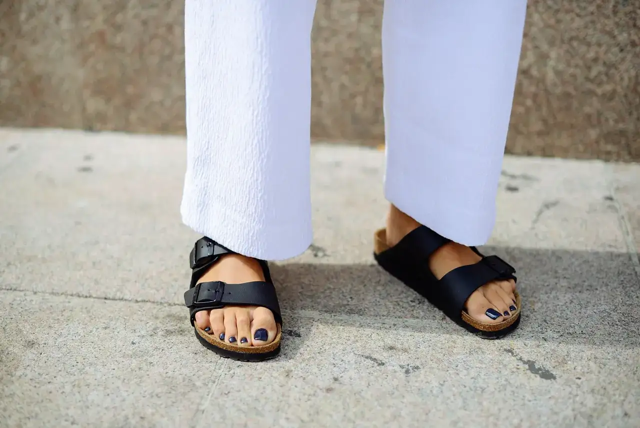 Pies con sandalias Birkenstock negras, ideales para aliviar la fascitis plantar. Pantalones blancos y u&ntilde;as oscuras.