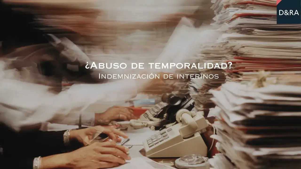 ¿Abuso de temporalidad? Indemnización de interinos tras 3 años. Escritorio caótico con pilas de papeles y manos trabajando.