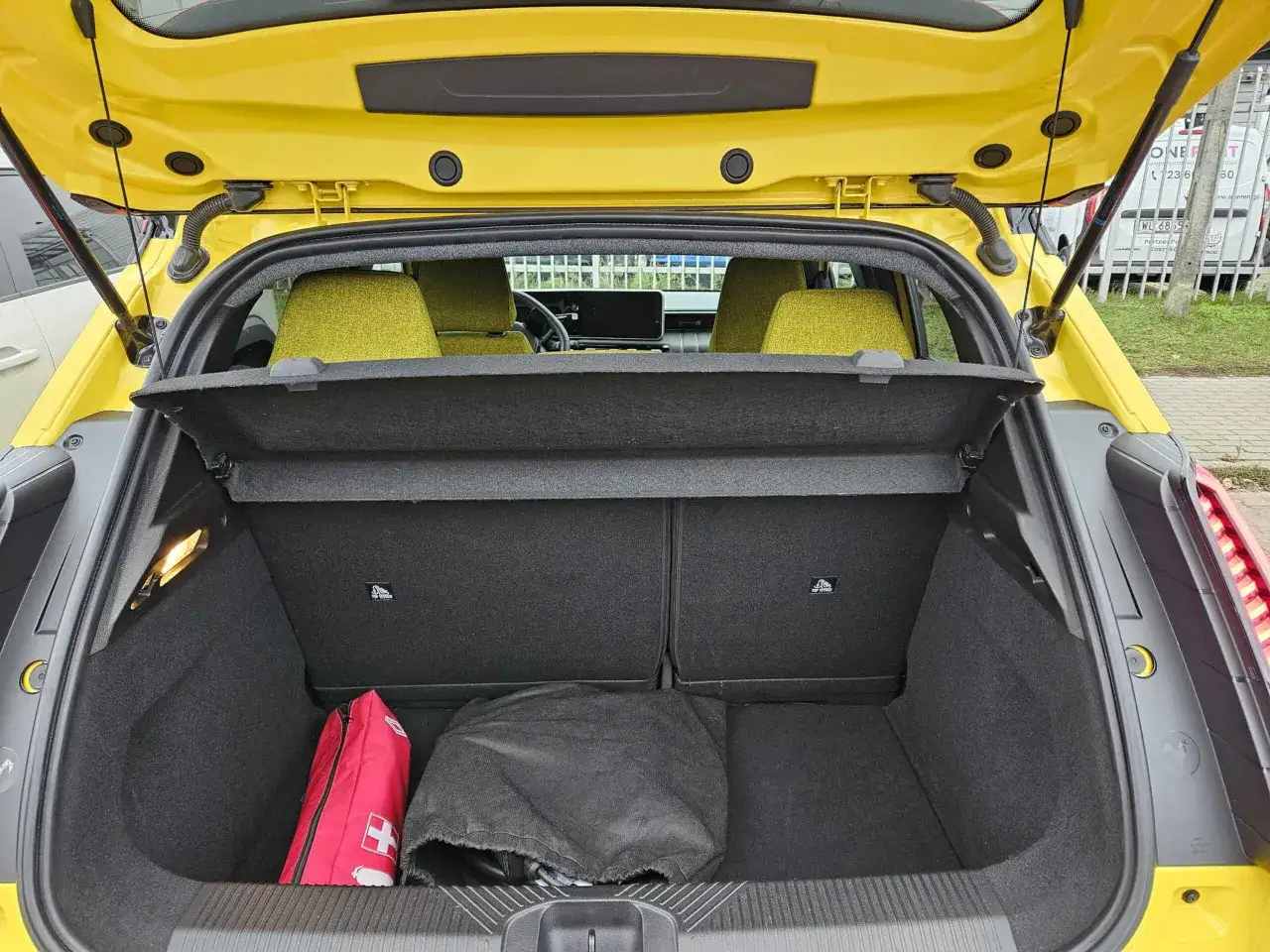 Bagażnik żółtego Renault 5 e-tech z apteczką i pokrowcem. Wnętrze auta z zielonymi siedzeniami i ekranem.