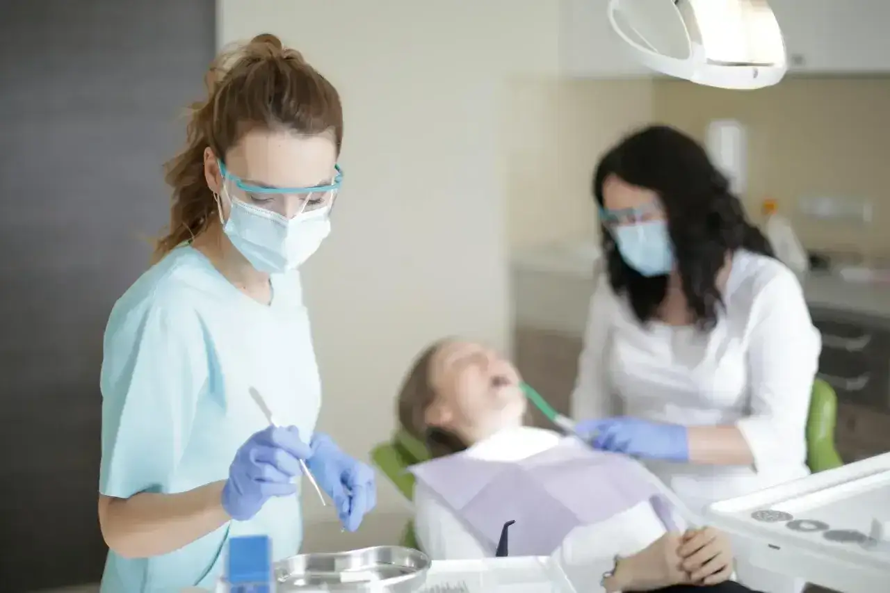 Dwie dentystki w maskach i okularach pracują przy pacjencie. Jedna z nich trzyma narzędzie, być może do usuwania pozostawionych szw&oacute;w nierozpuszczalnych.