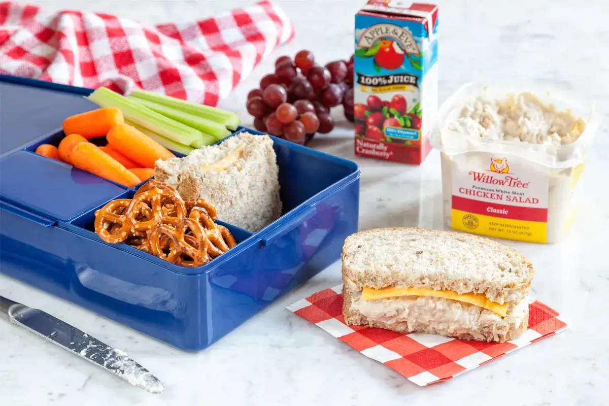 A blue bento box filled with a chicken salad sandwich, carrots, celery, and pretzels. A separate chicken salad sandwich and juice box are nearby.