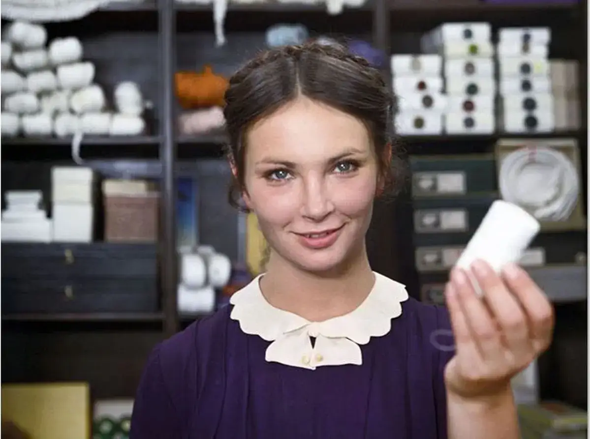 Anna Dymna w jednym z filmów, trzymająca biały przedmiot. W tle półki z materiałami.