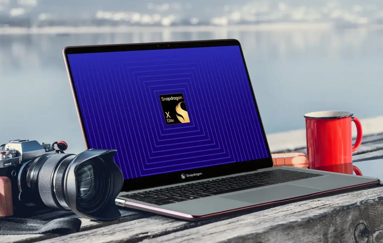 Ein Laptop mit Snapdragon X Elite Logo auf dem Bildschirm, daneben eine Kamera und eine rote Tasse am Seeufer.