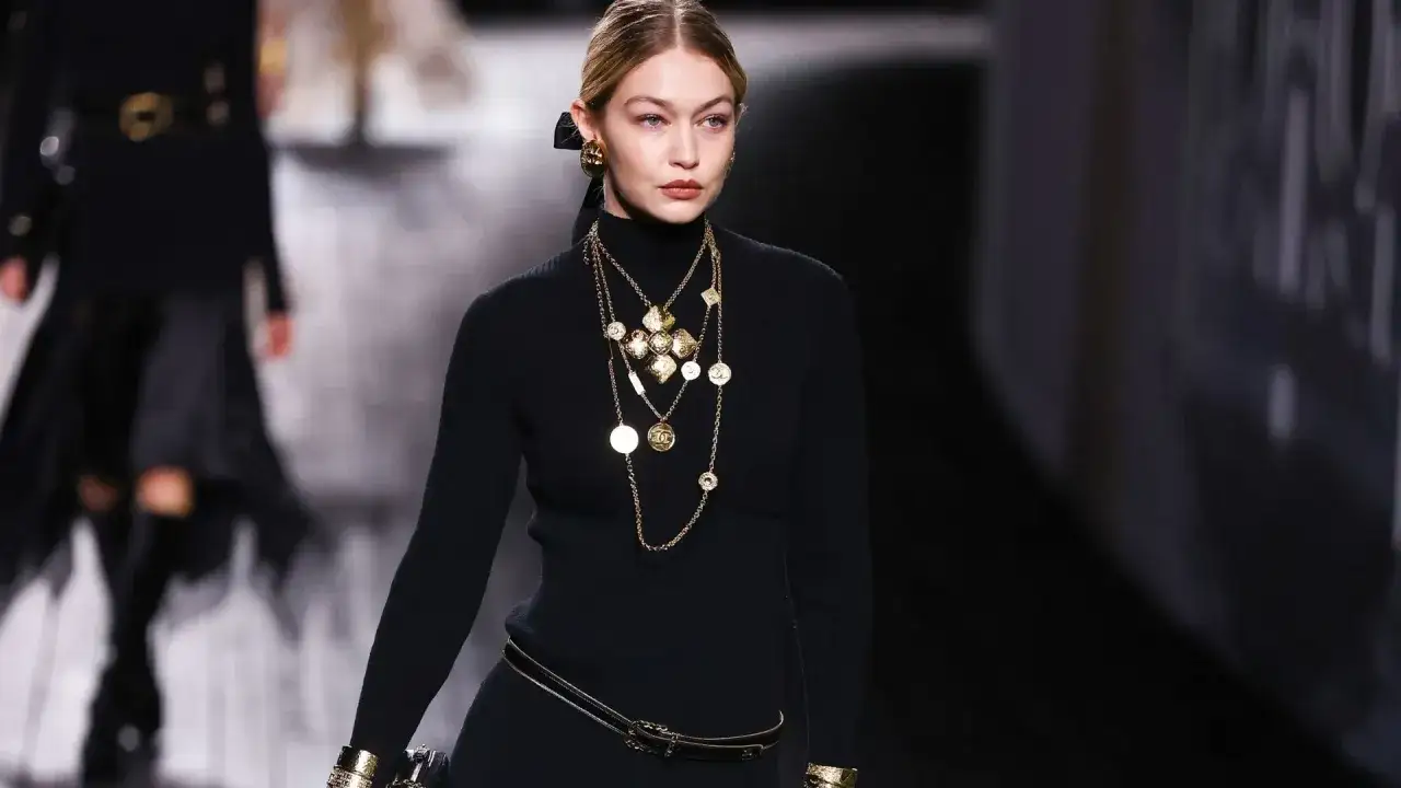 Modelka w czarnej sukience z golfem i bogatej biżuterii, nawiązującej do stylu moda lat 20. Złote naszyjniki i kolczyki dodają elegancji.