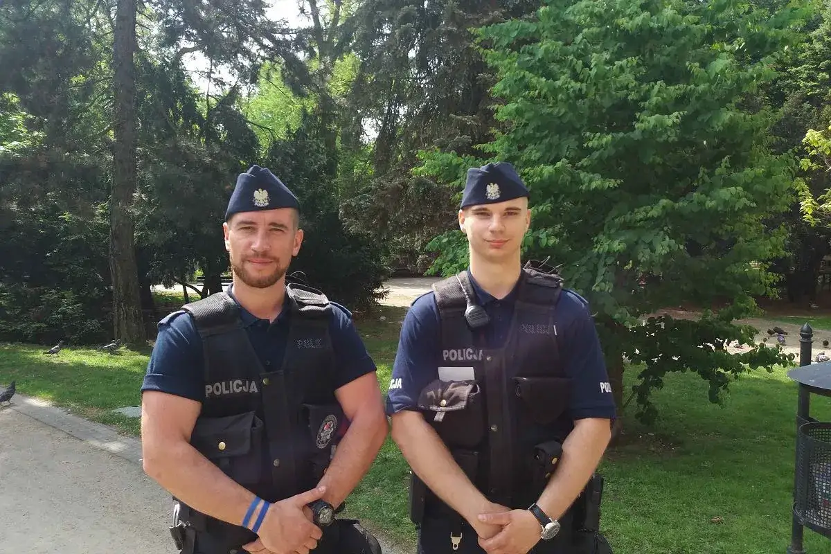 Dwóch policjantów w furażerkach, kamizelkach z napisem POLICJA, pozuje w parku.