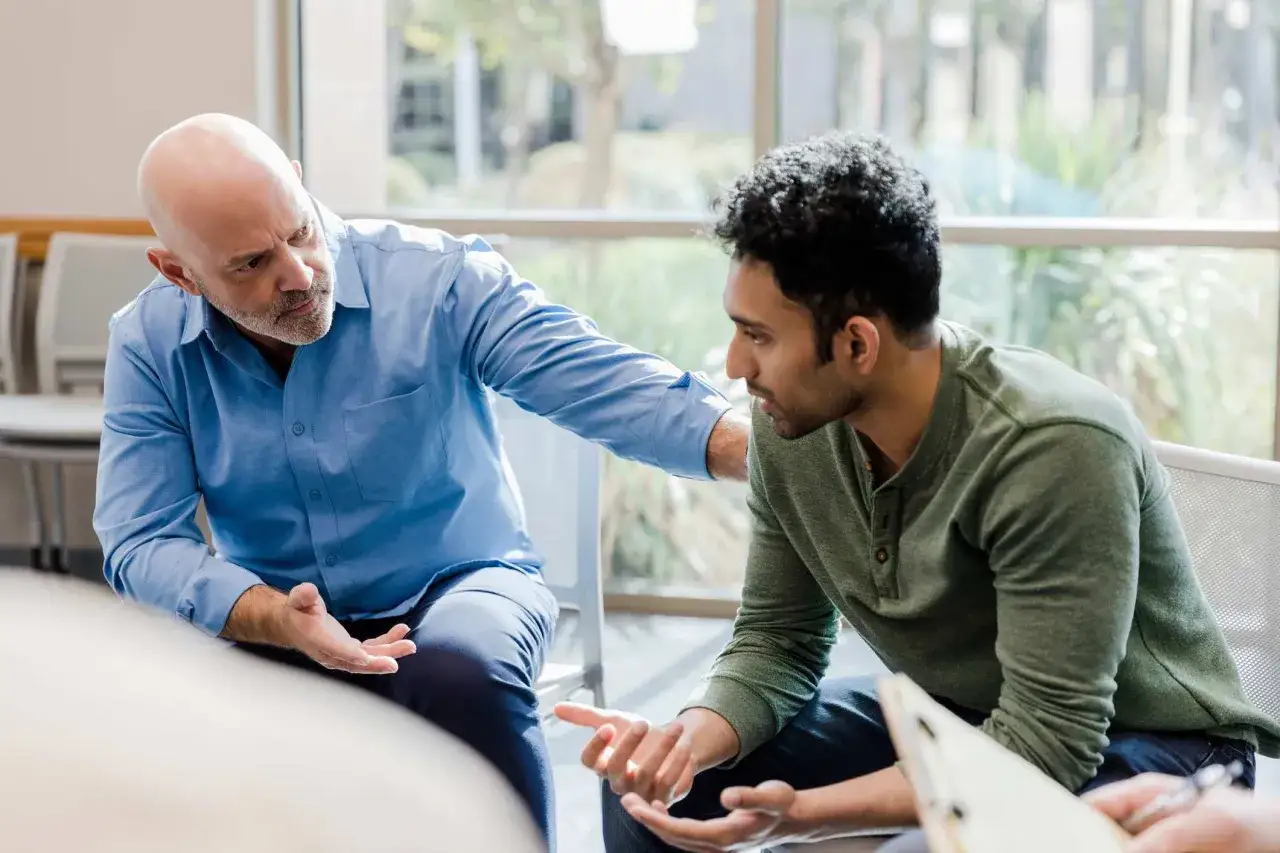 A man in a blue shirt places a hand on another man's shoulder, offering support. 