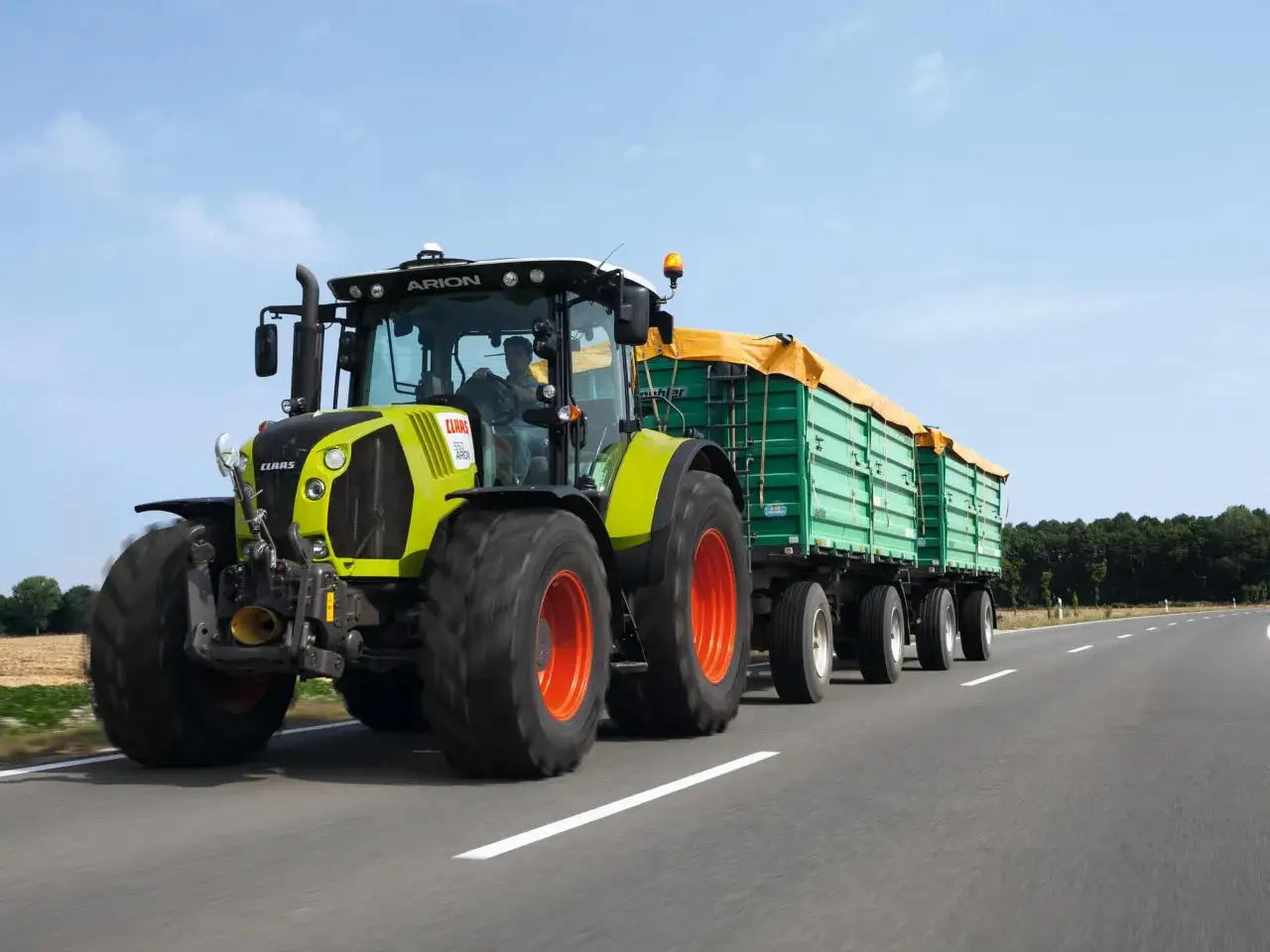 Ein Claas Arion Traktor mit Anhängern fährt auf der Straße. Ab wann darf man Traktor fahren? Mit 16 Jahren mit speziellem Führerschein.