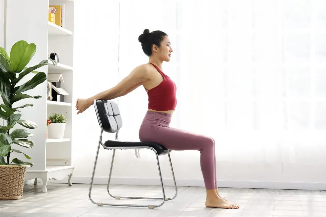 Femme pratiquant la gym douce Pilates, assise sur une chaise, bras derrière la tête pour un étirement.