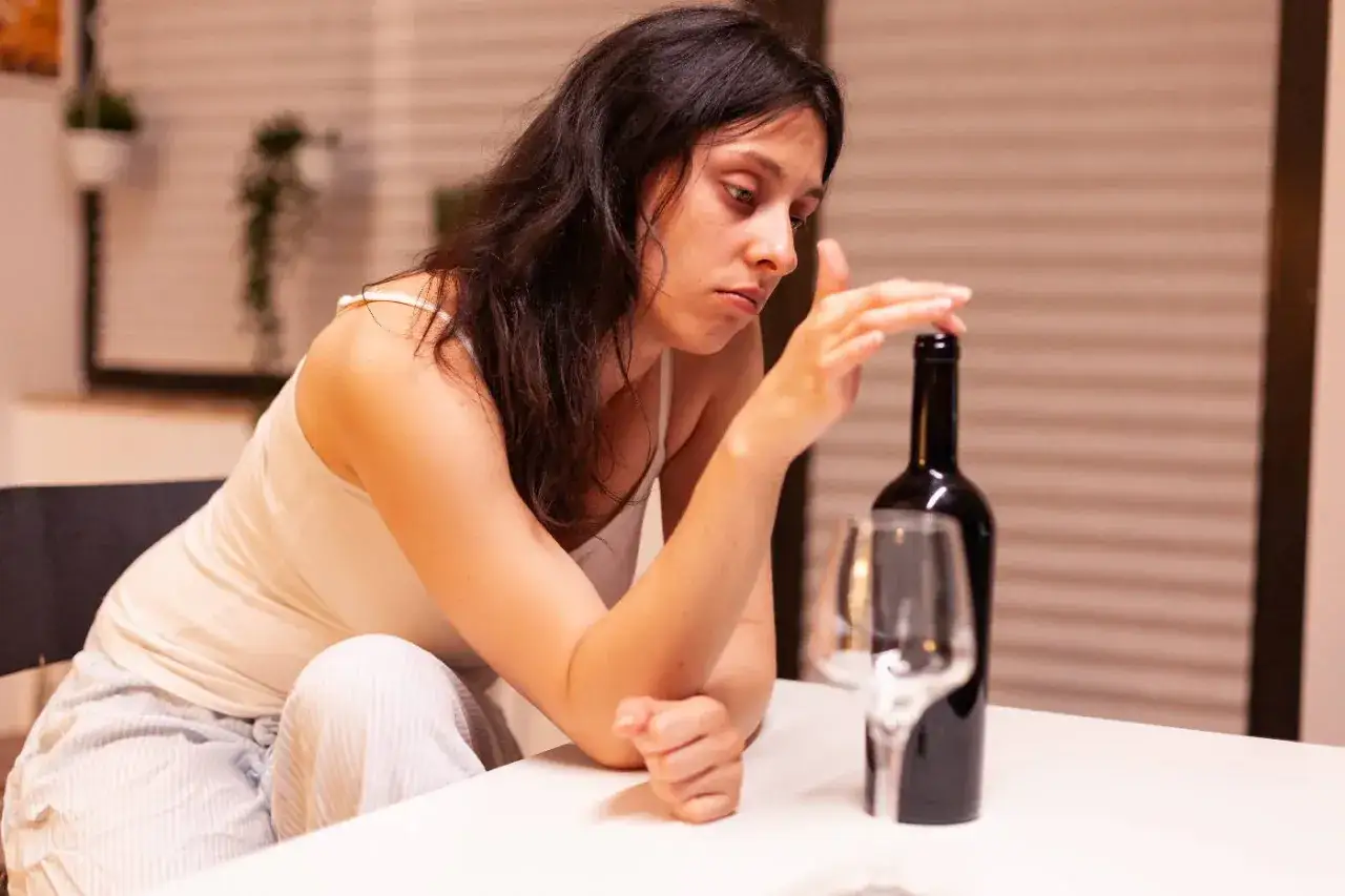 Zamyślona kobieta przy butelce wina, symbolizująca potrzebę skierowania na detoks alkoholowy.