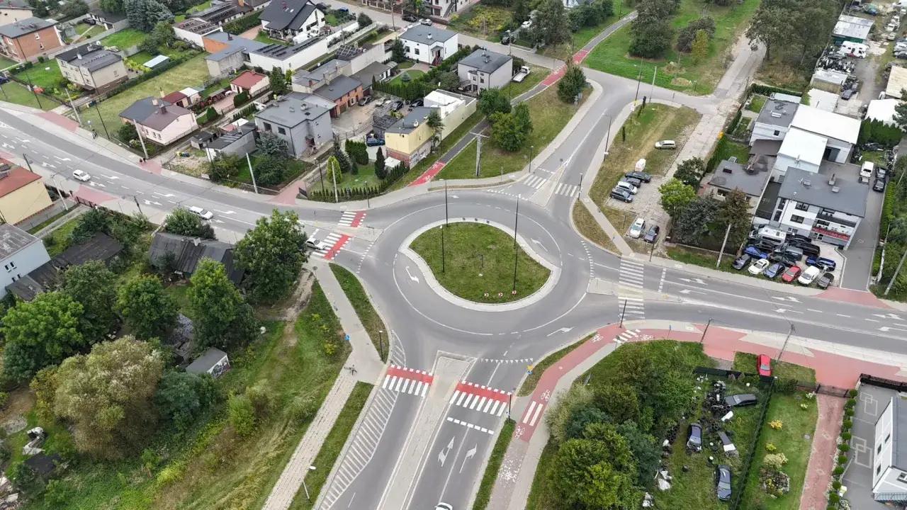 Rowerzysta na rondzie, otoczonym domami i samochodami.